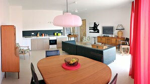 A modern kitchen with a dining area, featuring a round wooden table with a fruit bowl, a pink pendant light, and a TV on a wooden cabinet. - Large seaside penthouse, with breathtaking country views (Iż-Żebbuġ)