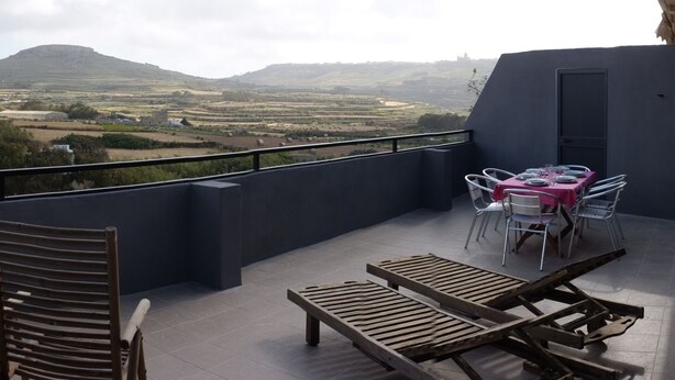 A balcony with a table set for dining, two wooden lounge chairs, and a view of a hilly landscape. - Large seaside penthouse, with breathtaking country views (Iż-Żebbuġ)