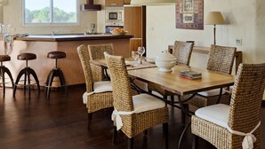 Un comedor espacioso con techo alto y vigas de madera, que incluye una mesa larga con sillas de mimbre y una isla central con taburetes de bar.