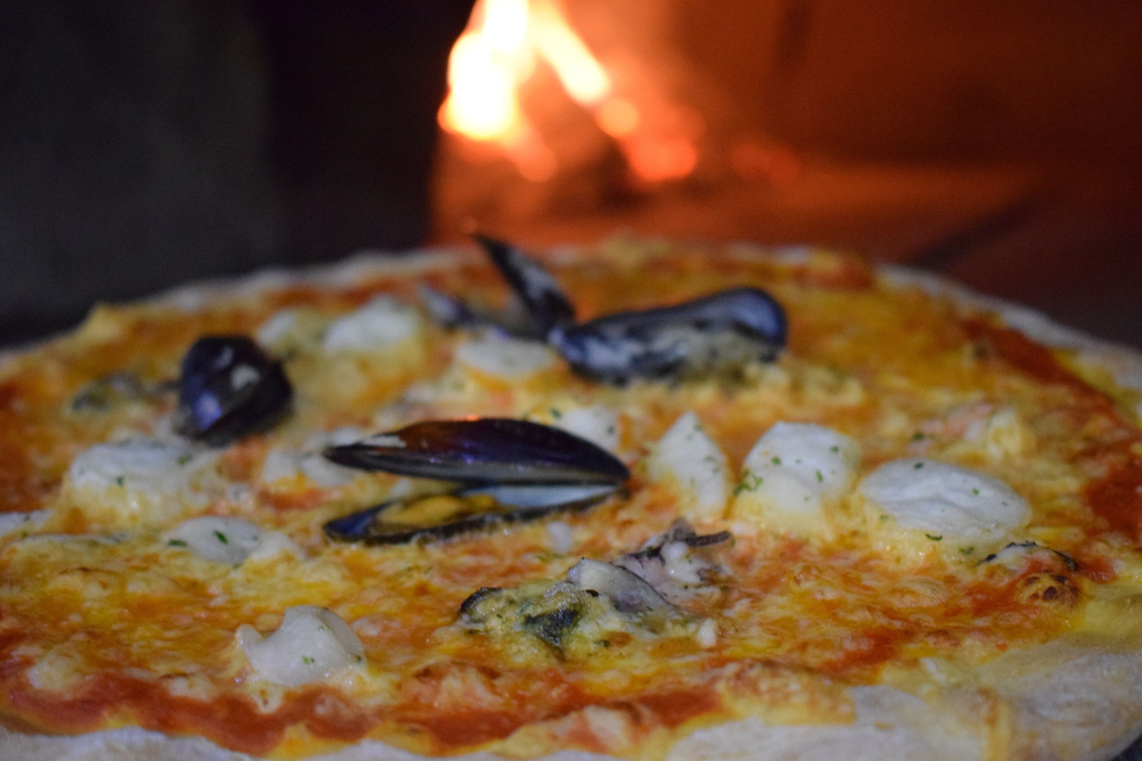 A pizza with seafood toppings, including mussels and fish, in front of a wood-fired oven.