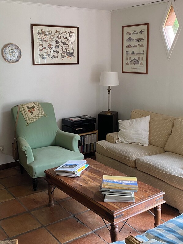 A living room with a green armchair, a beige sofa, a wooden coffee table with books, a lamp, and framed pictures on the wall.