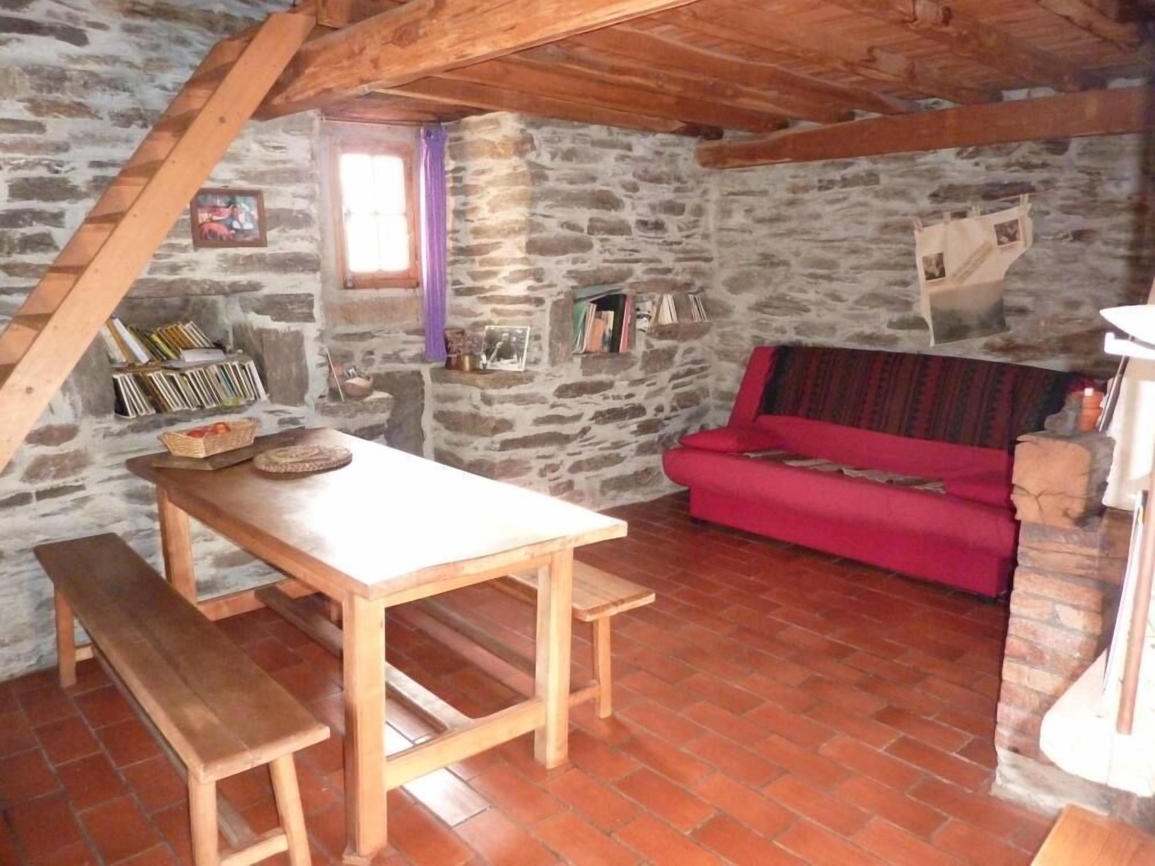 Una stanza rustica con parete in pietra, un tavolo di legno con panche, un divano rosso e una libreria piena di libri e oggetti decorativi.