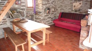 Una habitación rústica con pared de piedra, una mesa de madera con bancos, un sofá rojo y una estantería llena de libros y objetos decorativos.