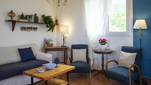 Un salon comprenant un canapé, deux fauteuils, une table basse en bois, une petite table d’appoint avec une lampe, une fenêtre avec des rideaux et une étagère avec des livres et des objets décoratifs.