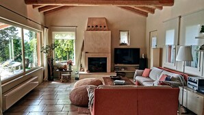 Una sala de estar acogedora con chimenea, un sofá, un televisor y un amplio ventanal con vistas a un jardín.