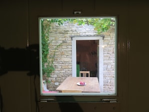 Une fenêtre donnant sur un mur de pierre et des lianes vertes, une table en bois avec une assiette de fruits et une chaise.