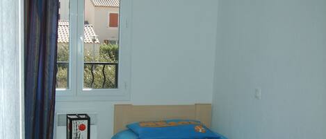 A bedroom with a bed, a small table, a window with curtains, a blue hanging light, and a tiled floor.