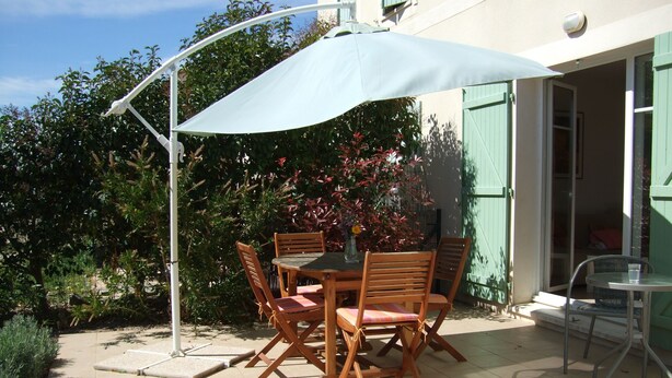 Une terrasse extérieure avec une table et des chaises, un parasol blanc et une vue sur un bâtiment aux volets verts.
