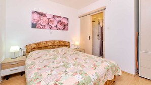 Ein Schlafzimmer mit floralem Bettüberwurf, einem Nachttisch mit Lampe und einem sichtbaren Duschbereich durch eine geöffnete Tür.