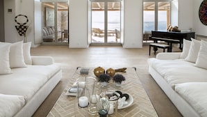 Ein geräumiges Wohnzimmer mit einem Klavier, weißen Sofas und Meerblick durch große Glasfenster.