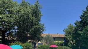 Un espace piscine agrémenté de chaises longues blanches et de parasols rouges, entouré d’arbres et d’un bâtiment doté d’un toit en tuiles.