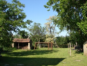 A grassy outdoor area with a wooden swing set, a small brick structure, and trees. - Serbian Village Farmhouse (Stara Moravica)