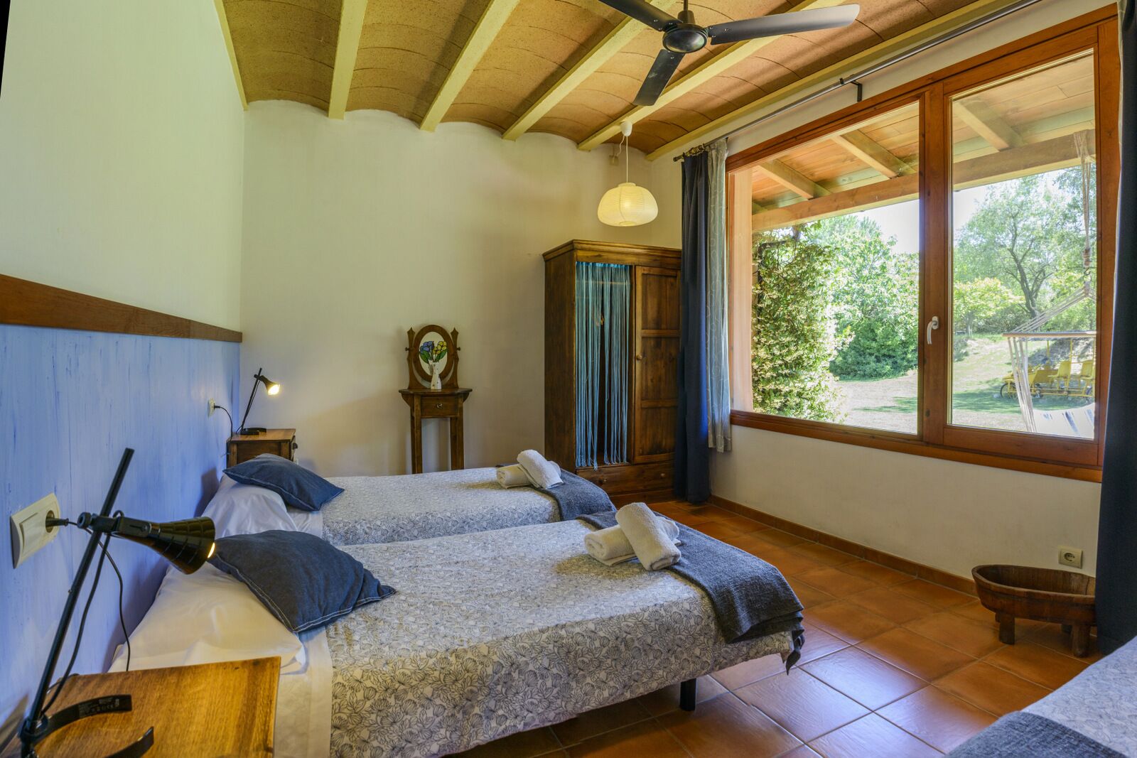 Une chambre avec deux lits, des poutres en bois au plafond, une grande fenêtre donnant sur la verdure et un meuble en bois.