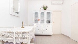 A white room with a bed, a white table with a lace tablecloth, a white cabinet with glass doors, and a white air conditioning unit on the wall.
