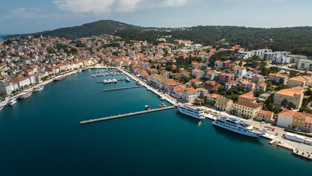 An aerial view of a coastal town with a marina, buildings, and a hill in the background.