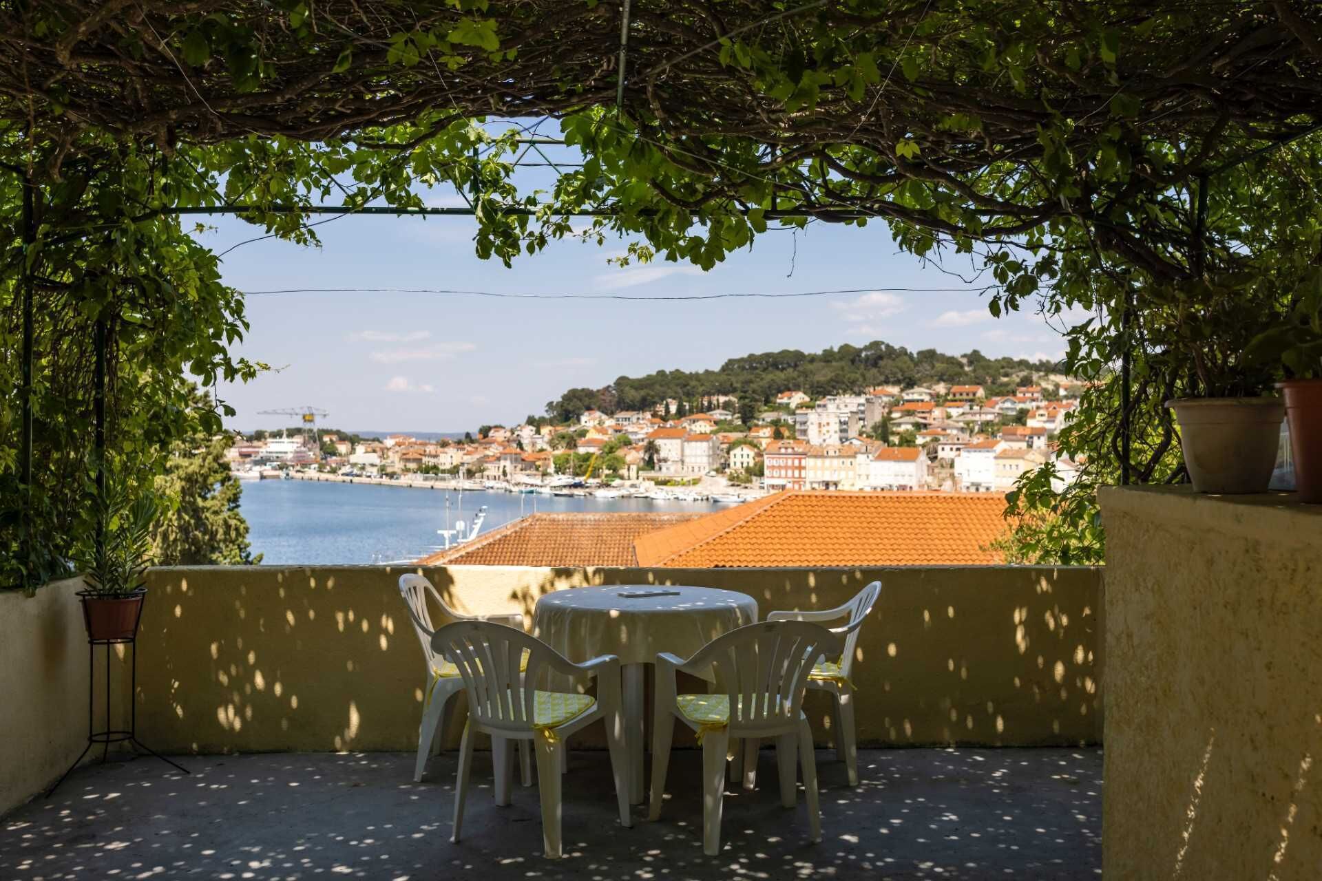 An outdoor patio with a table and chairs, overlooking a coastal town with buildings and boats.