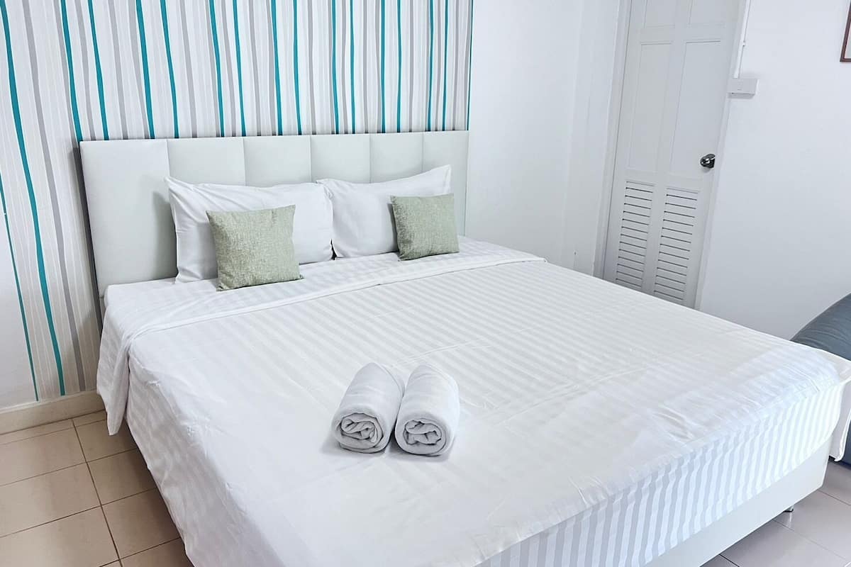 A neatly made bed with white linens and two rolled towels, a white headboard, and a striped curtain in the background.