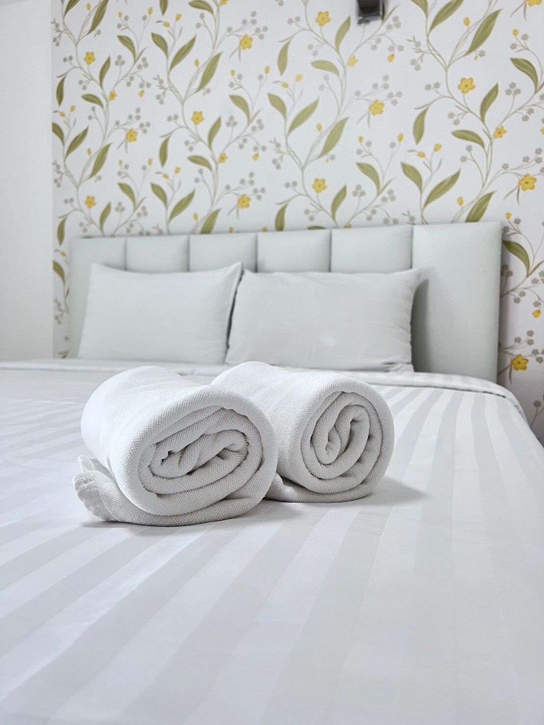 Ein ordentlich bezogenes Bett mit aufgerollten weißen Handtüchern, einem hellen Kopfseitenverkleidungen und blumigem Wandpapier.