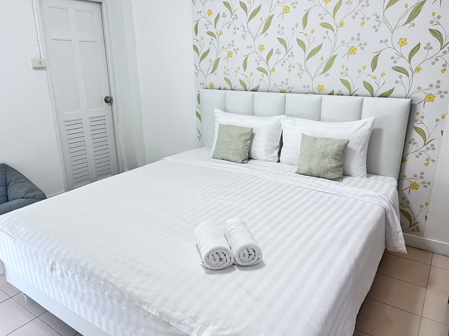 A neatly made bed with white linens and two rolled towels, a white headboard, and a floral wallpaper pattern.