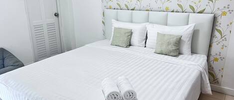 A neatly made bed with white linens and two rolled towels, a white headboard, and a floral wallpaper pattern.