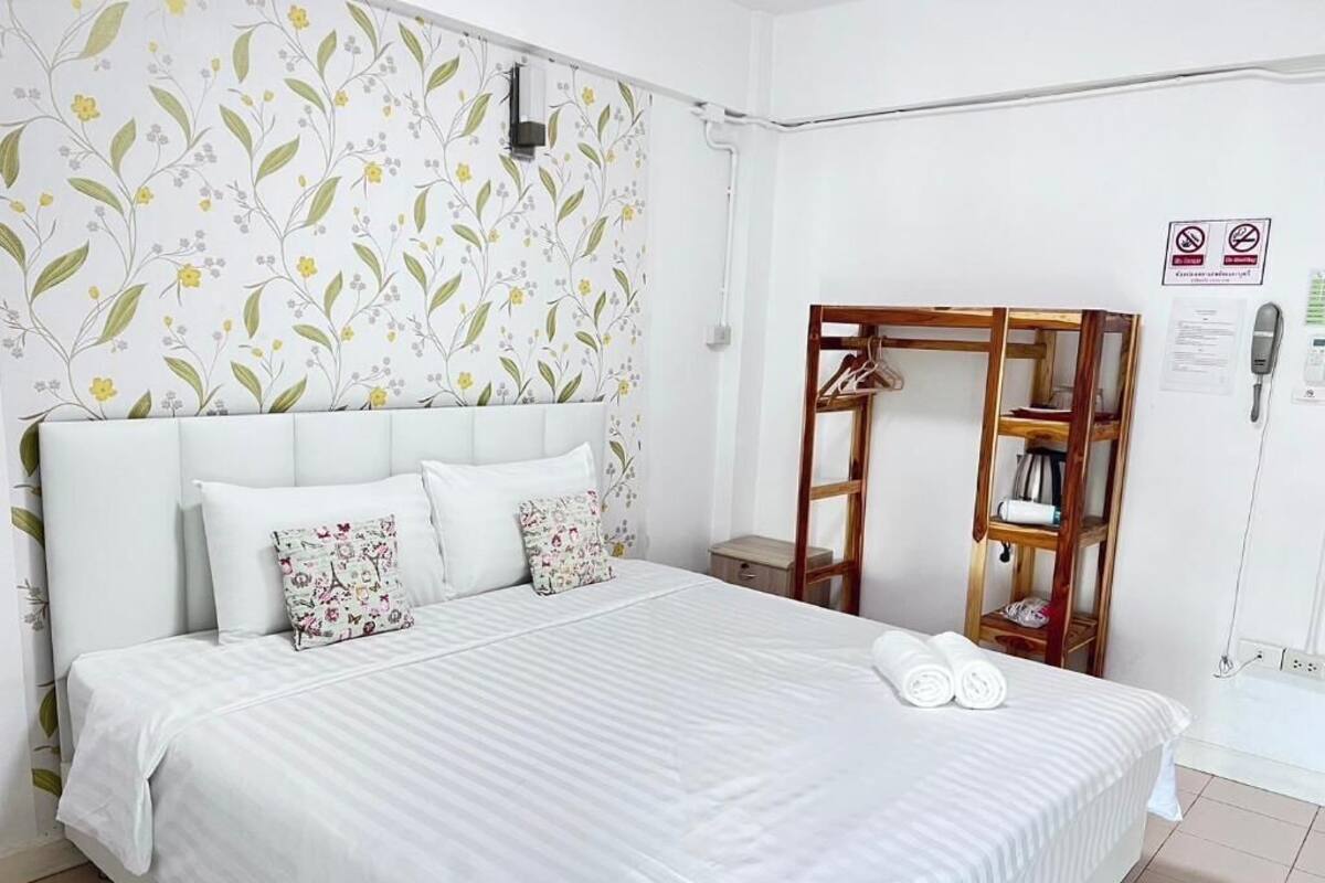 A neatly made bed with white bedding, a floral-patterned pillow, and a wooden hanger stand with clothes.