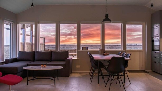 A modern living room with a large window overlooking a scenic view, a dining area with chairs, and a kitchen area. - Bryggjur (Rangárþing eystra)