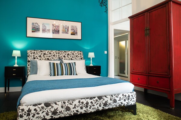 A bedroom with a teal wall, a bed with floral-patterned bedding, two bedside tables with lamps, and a red cabinet.