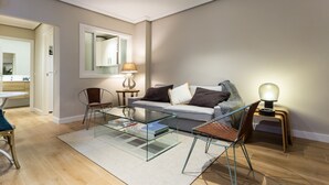 A modern living room with a glass coffee table, a sofa with cushions, a side table with a lamp, and a chair.