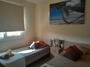 A bedroom with a single bed, a bedside table, a nightstand, a window, and a wall-mounted painting of a beach scene.