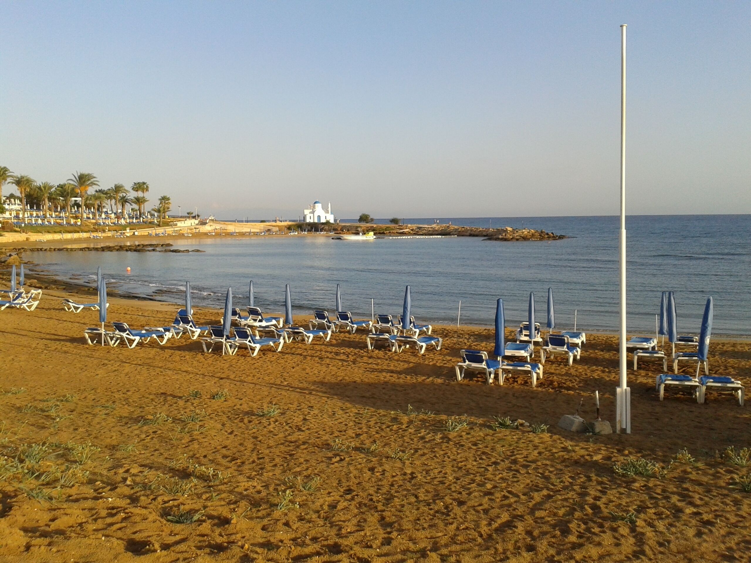 Une plage bordée de rangées de chaises longues et de parasols, un bâtiment blanc surmonté d’un dôme, et un ciel dégagé.