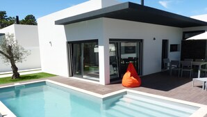 Een modern zwembadterras met een helderblauw zwembad, een eigentijds gebouw met grote glazen deuren en een zitgedeelte met stoelen en een tafel.