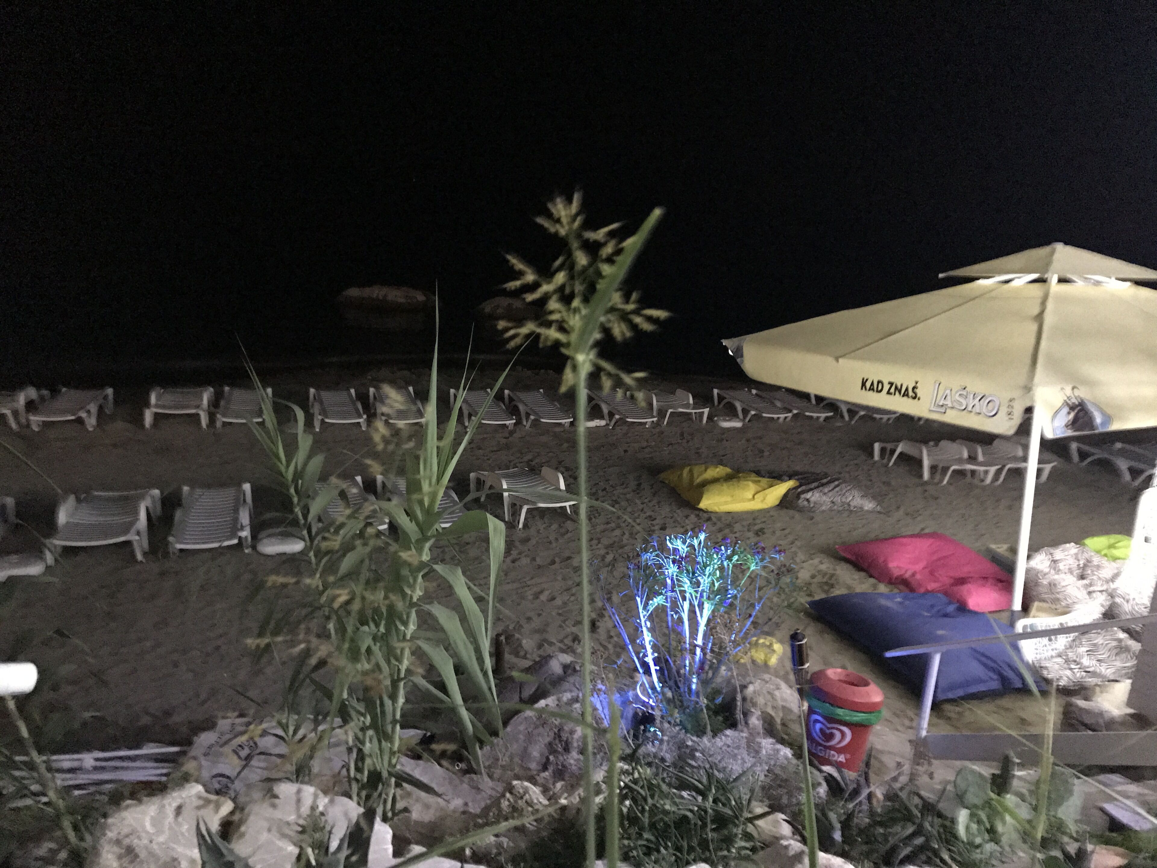 Un área de descanso junto a la playa con filas de tumbonas, una sombrilla grande y algunas luces decorativas.
