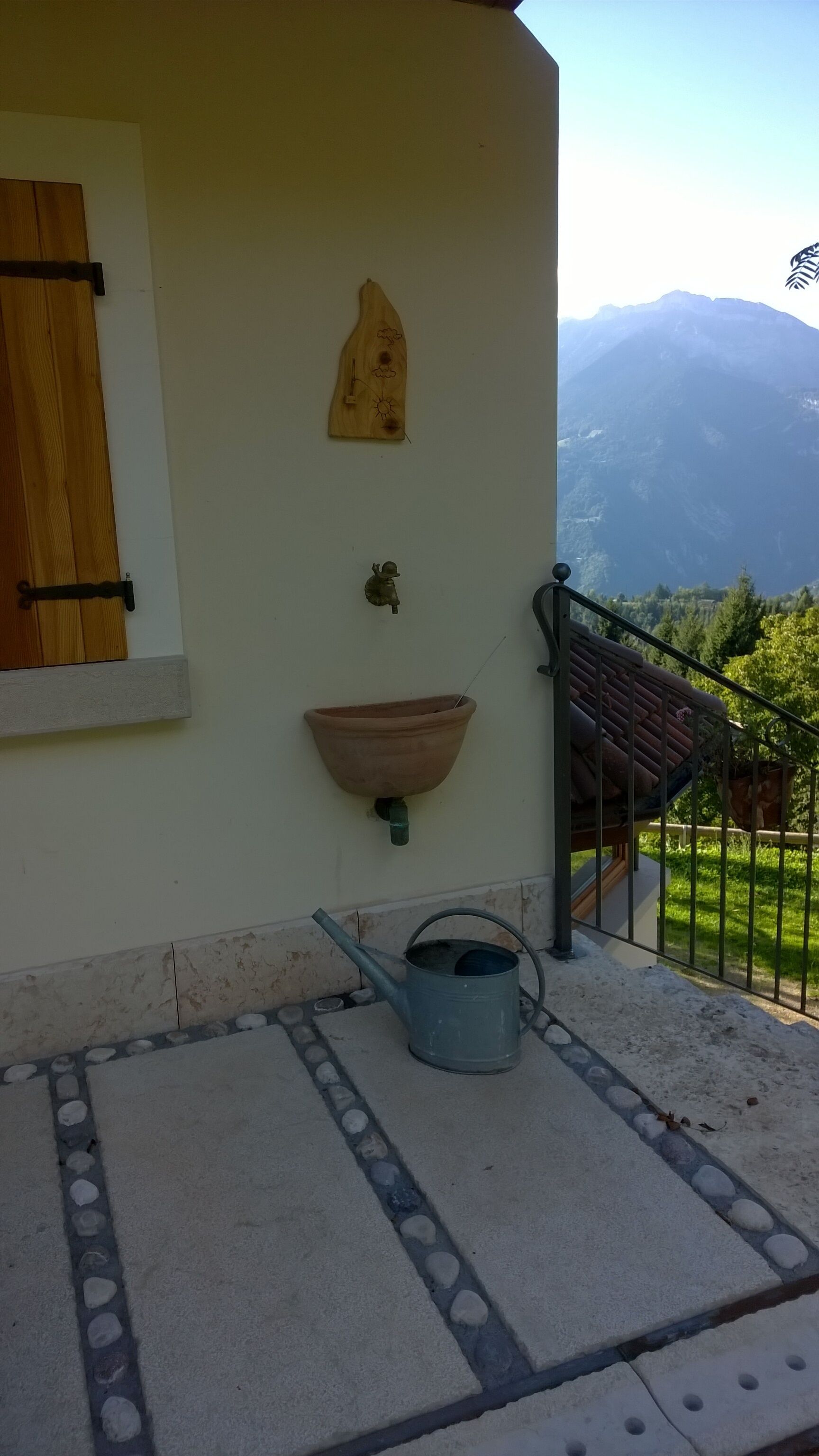 Un balcón con vistas a las montañas, un adorno de pared de madera, una maceta de barro y un regador de metal.
