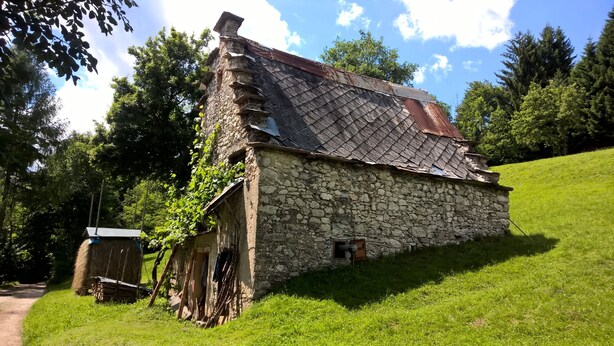 A rustic stone building with a sloped roof, surrounded by lush greenery and a clear sky.