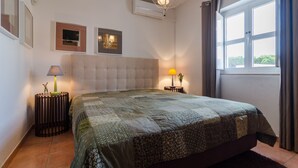 Ein Schlafzimmer mit einem großen Bett, zwei Nachttischlampen, einem fensterbelaubten Fenster und Wand-Klimaanlage.