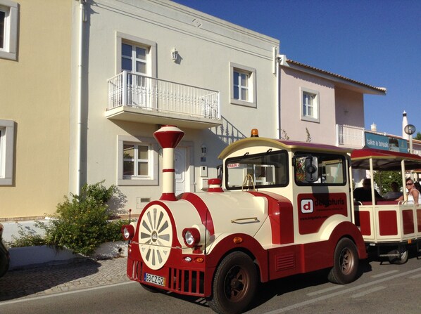 A vintage red and white train with a front funnel and a bell, parked in front of a building with balconies and windows.