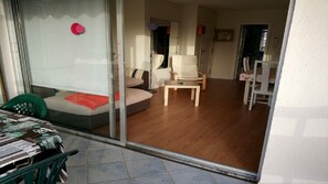 A room with a sliding glass door leading to a balcony, a sofa with a red blanket, a wooden coffee table, and a chair.
