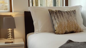 Una cama bien hecha con sábanas blancas y un cojín estampado, una mesita de noche con una lámpara, y un cuadro en la pared.