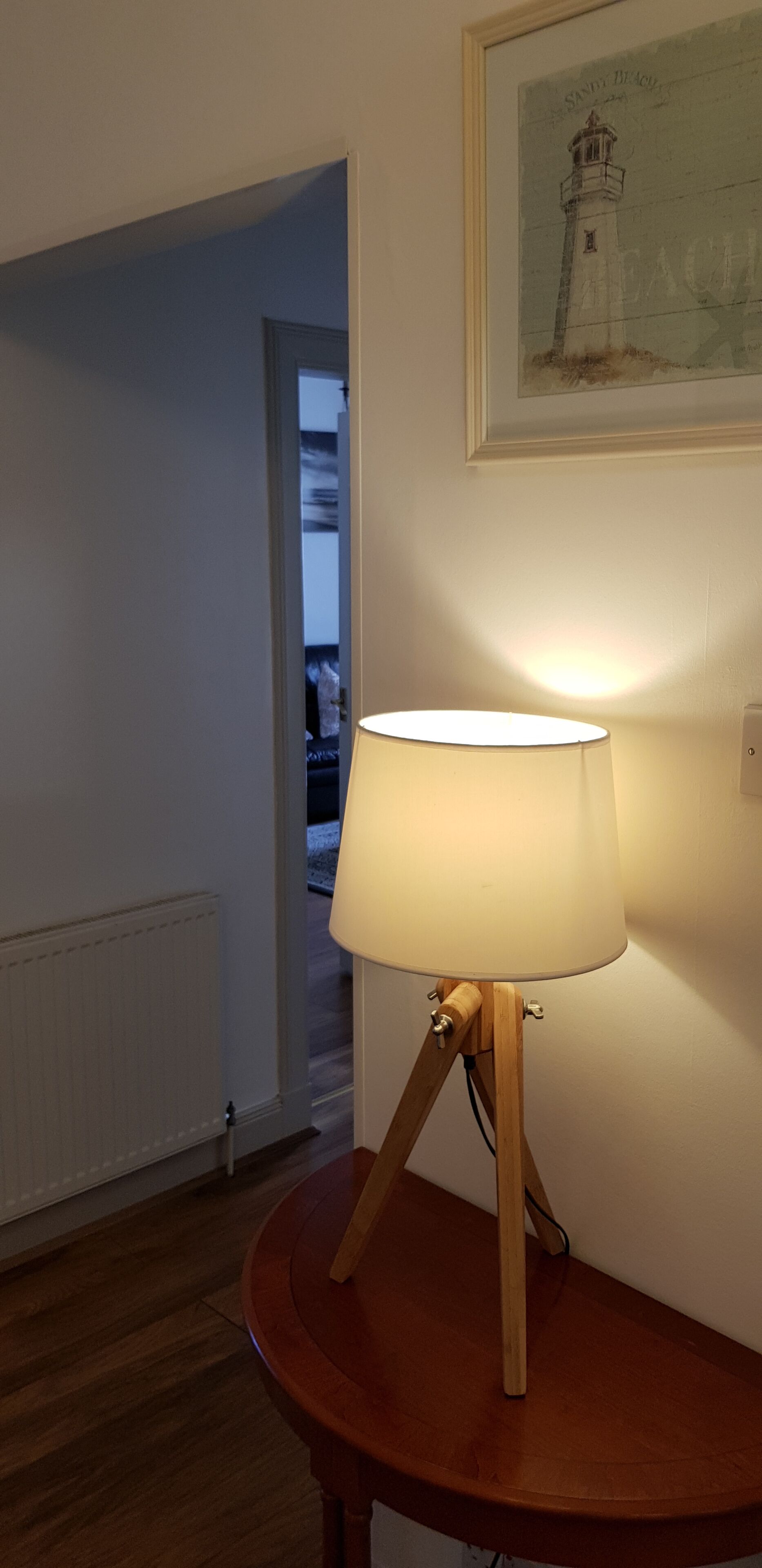 A room with a wooden table lamp on a round table, a framed picture on the wall, and a doorway leading to another room.