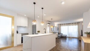 Una cocina moderna con armarios blancos, una isla de mármol, iluminación colgante y un comedor con sofá y mesa.