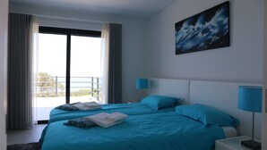 Une chambre d’hôtel avec deux lits, une grande fenêtre avec des rideaux, un tableau au mur et une table de chevet avec une lampe.