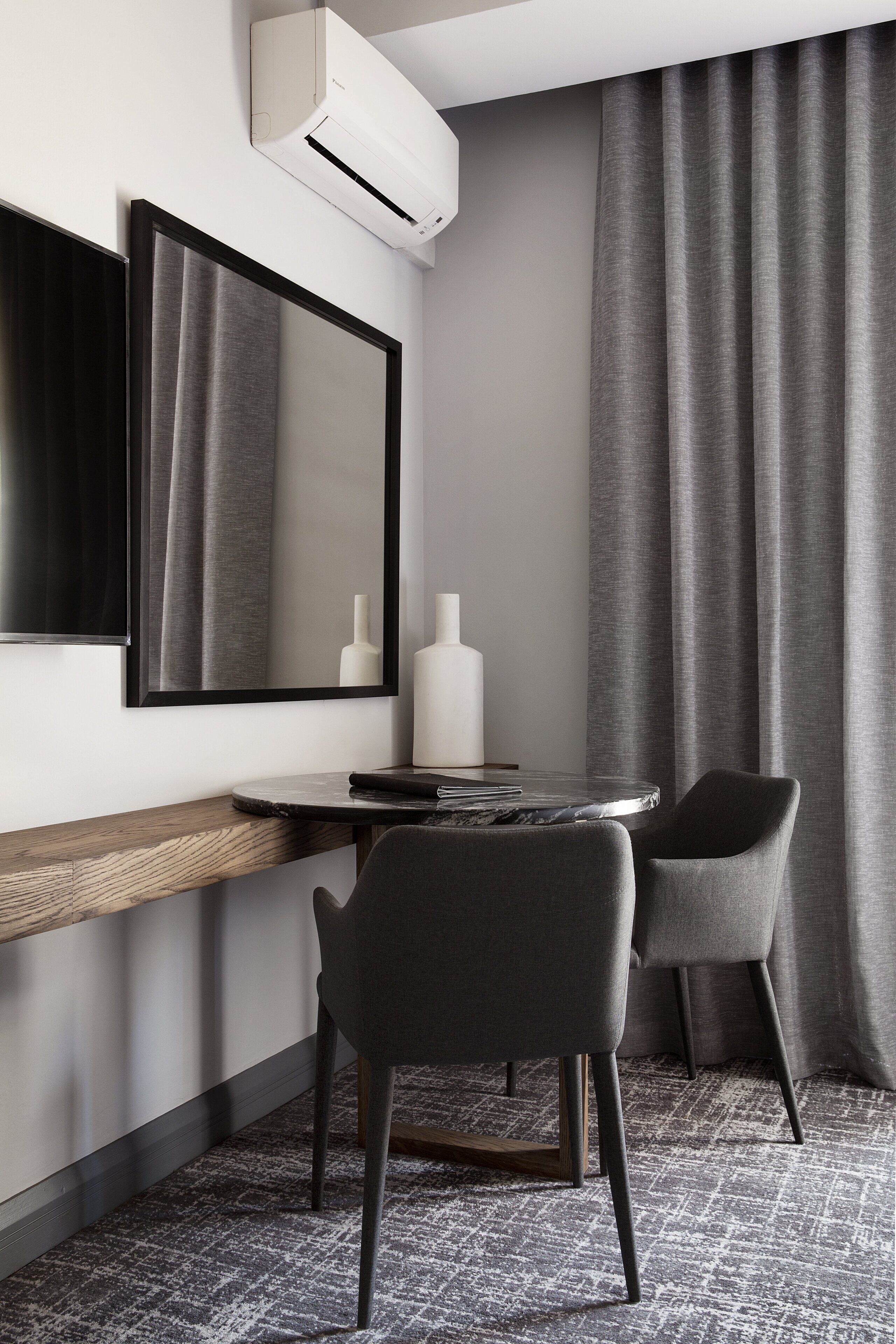 A modern hotel room with a large mirror, a round table, two chairs, a flat-screen TV, and an air conditioning unit.