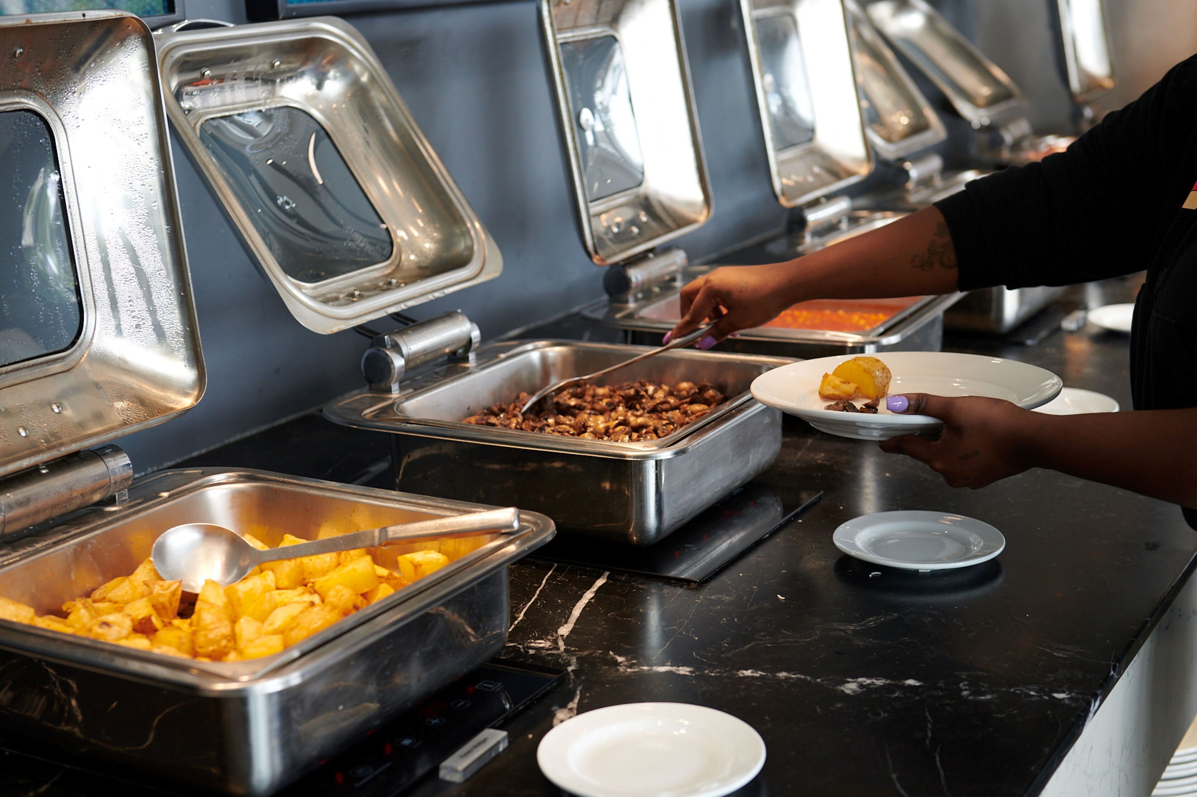 A buffet setup with various food items in metal containers, a person serving themselves, and a spoon in one of the containers.