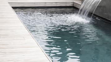 Una piscina con cascada, sillas de descanso y un cartel con una 'C' en una pared de azulejos.