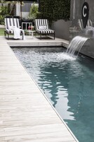 Una piscina con cascada, sillas de descanso y un cartel con una 'C' en una pared de azulejos.