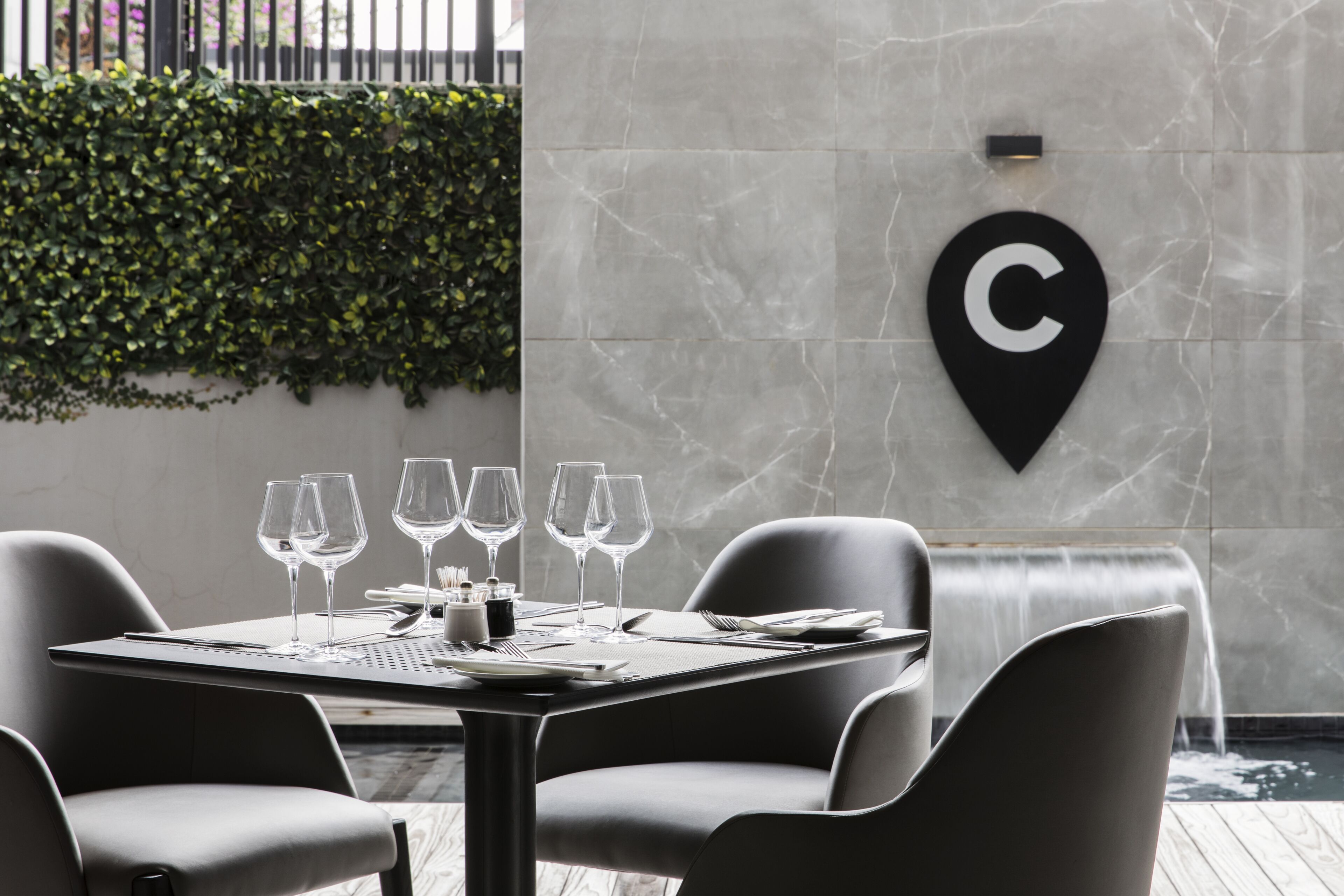 A dining area with a table set for four, a black wall-mounted sign with a 'C', and a green hedge in the background.