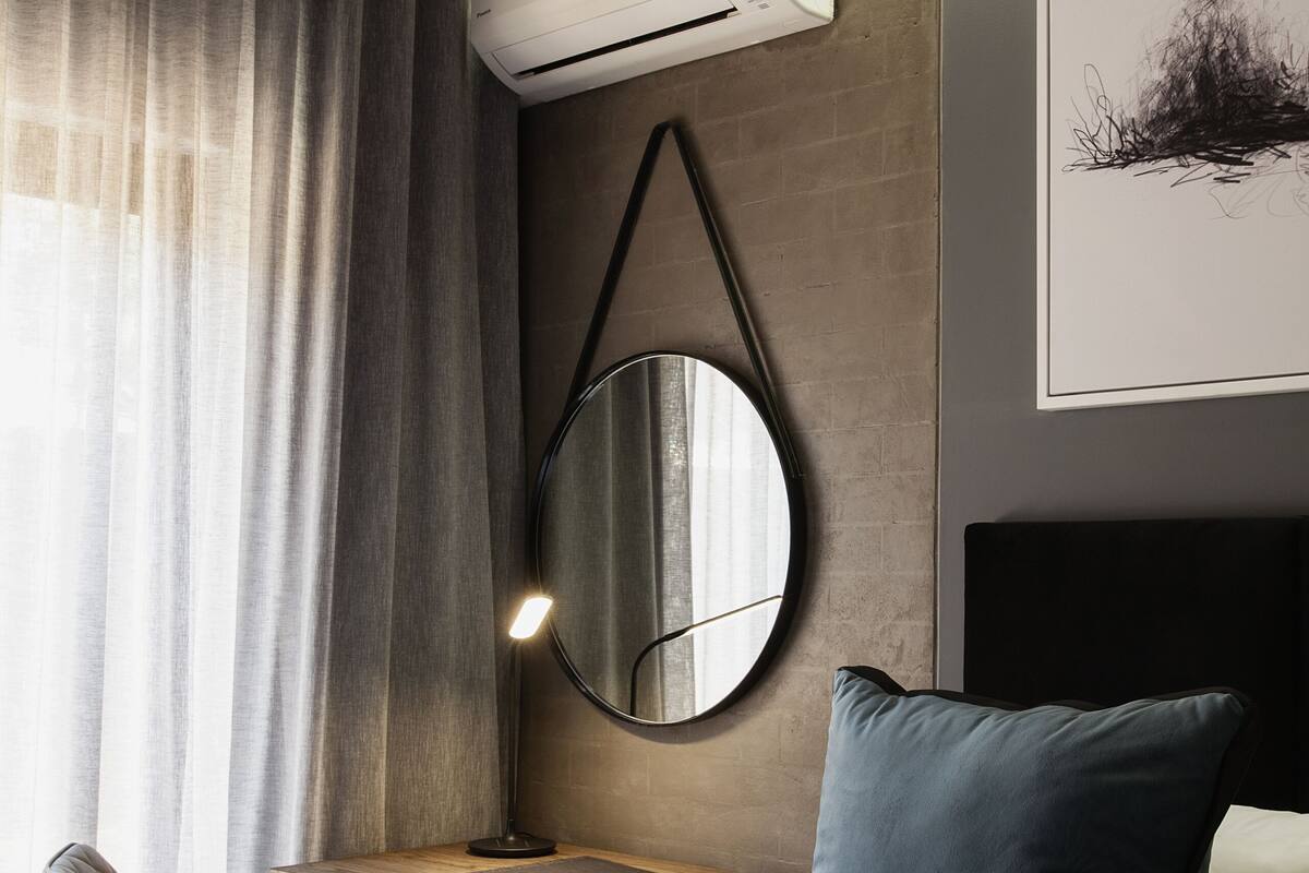 Een hotelkamer met een bed, een bureau met stoel, een ronde spiegel, een airconditioner en een kunstwerk in een lijst aan de muur.