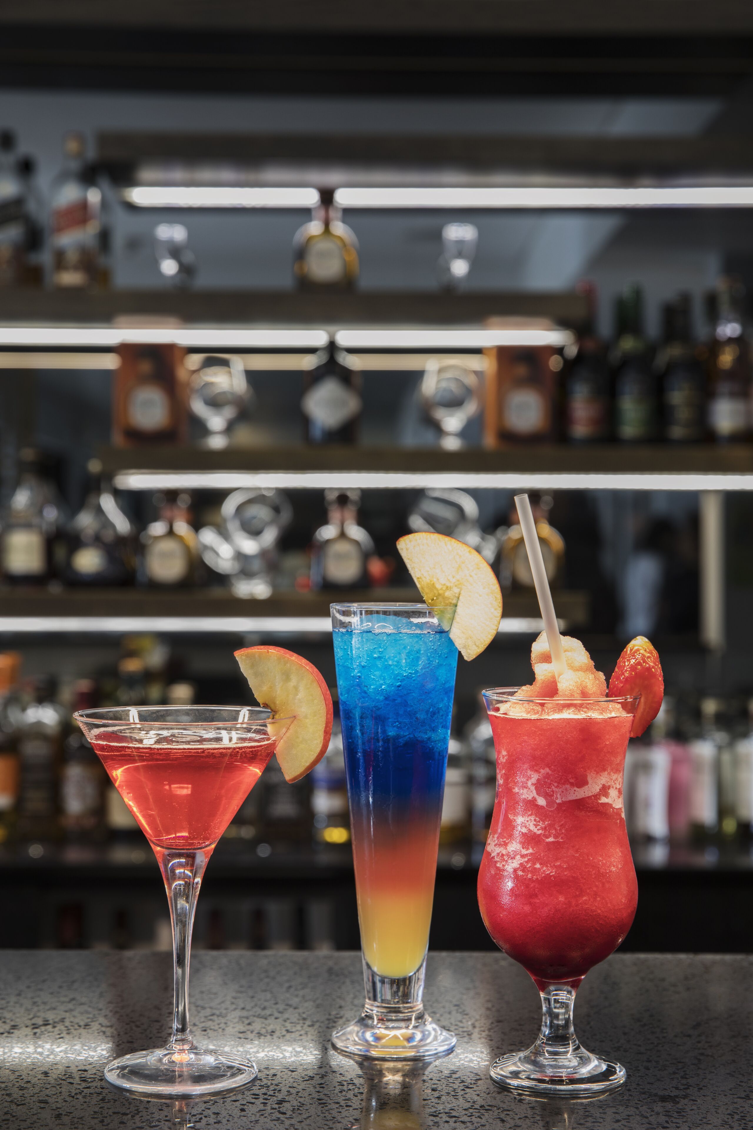 Three cocktails on a bar counter with shelves of bottles in the background.