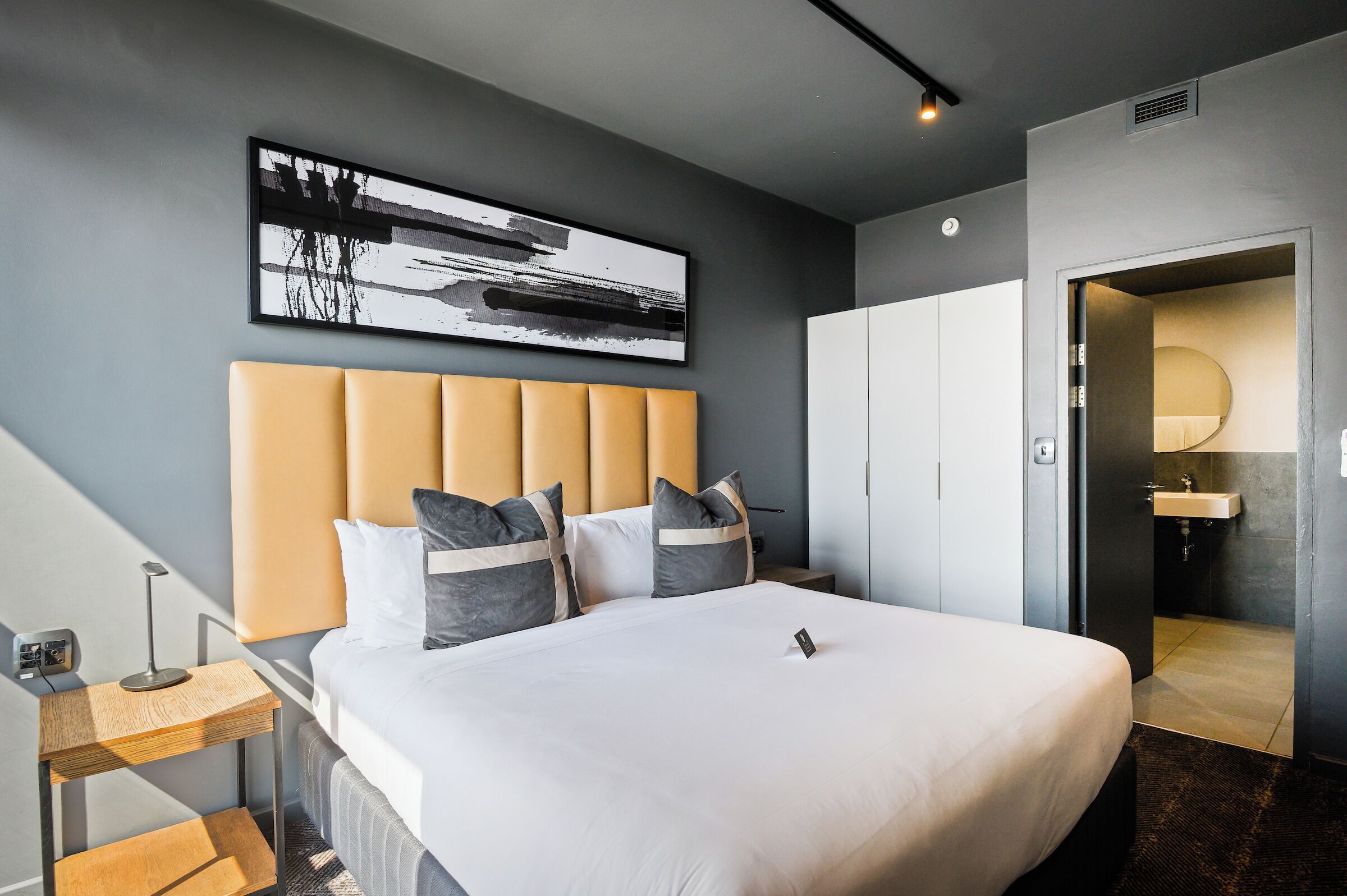 A modern hotel room with a large bed, a bedside table with a lamp, a wall-mounted artwork, and a view into the bathroom.