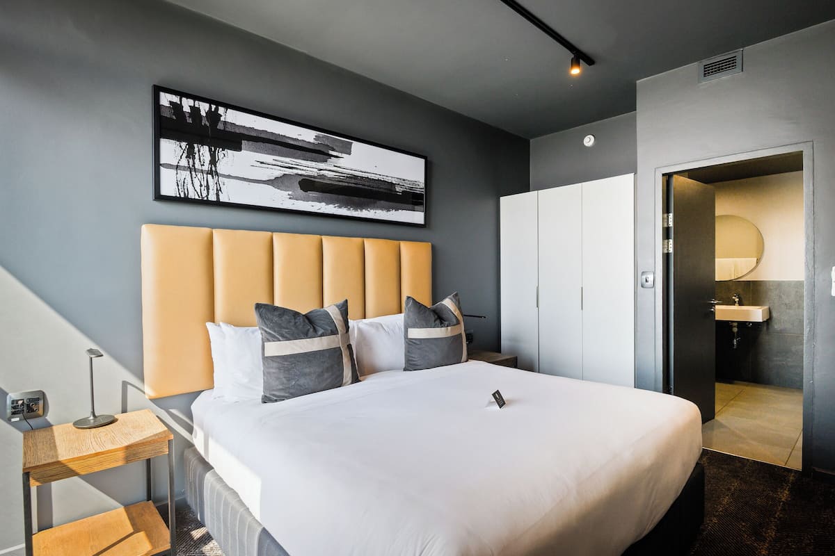 A modern hotel room with a large bed, a bedside table with a lamp, a wall-mounted artwork, and a view into the bathroom.